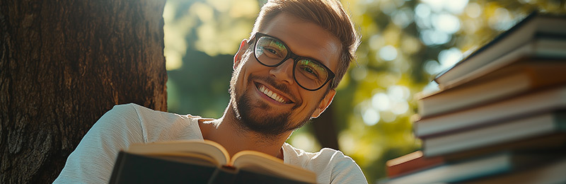Ein junger Mann liest lächelnd ein Buch unter einem Baum mit einem ganzen Stapel Bücher vor sich